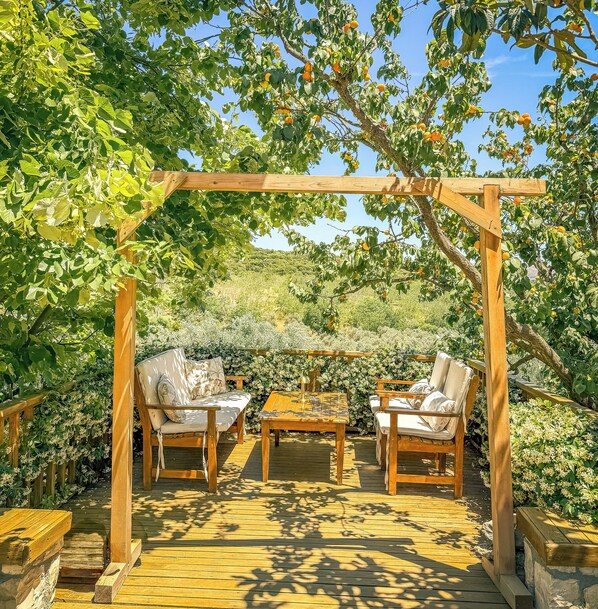 An outdoor wooden pergola with a table and two chairs underneath, surrounded by lush greenery and fruit trees.