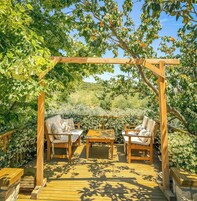 An outdoor wooden pergola with a table and two chairs underneath, surrounded by lush greenery and fruit trees.