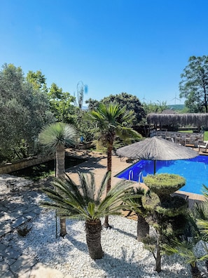 A pool area with a clear blue pool, surrounded by palm trees and a thatched umbrella. - Casa Del Olivia (Seferihisar)