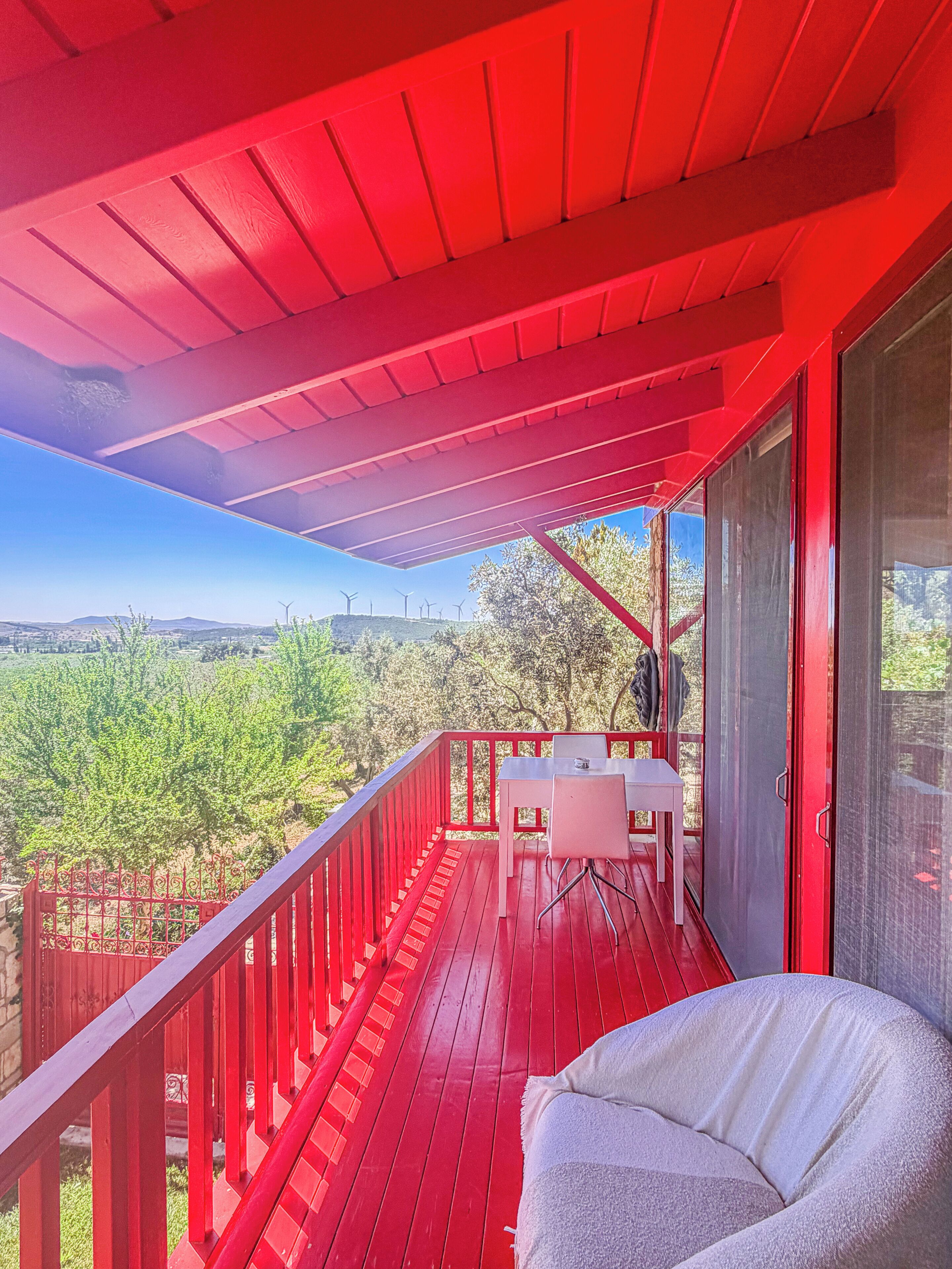 A red balcony with a table and chairs, overlooking a green landscape with wind turbines in the distance.
