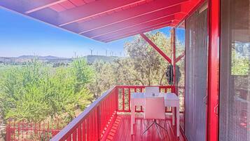 A red balcony with a table and chairs, overlooking a green landscape with wind turbines in the distance.