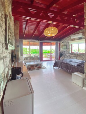 A room with a stone wall, a bed with a dark bedspread, a white sofa, a coffee machine, and a large window with a view of greenery. - Casa Del Olivia (Seferihisar)