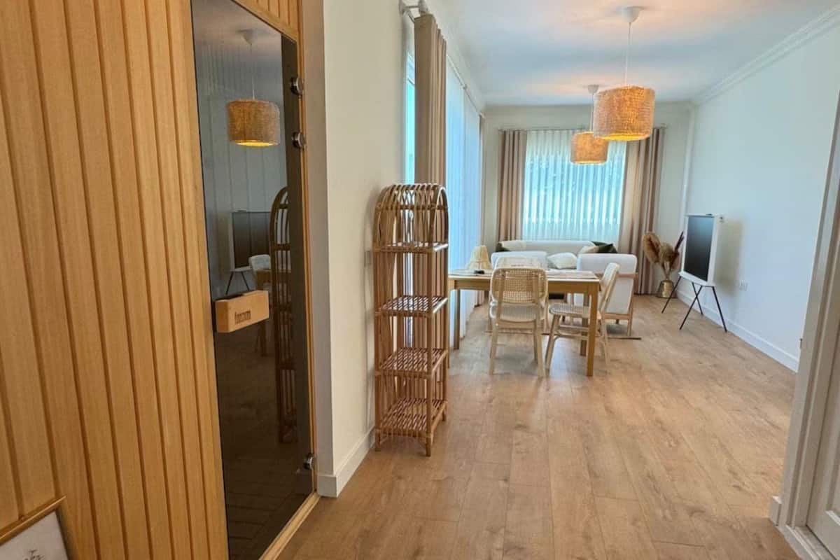 A spacious room with a wooden floor, a dining area with a table and chairs, a wicker chair, and a large mirror.