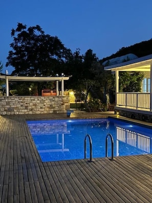 Une piscine dotée d’une terrasse en bois, d’un mur en pierre et d’un espace couvert éclairé.