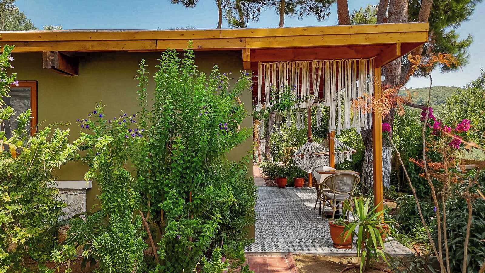 Un espacio de estar al aire libre con techo, que incluye una mesa y sillas, rodeado de abundante vegetación y plantas en macetas.
