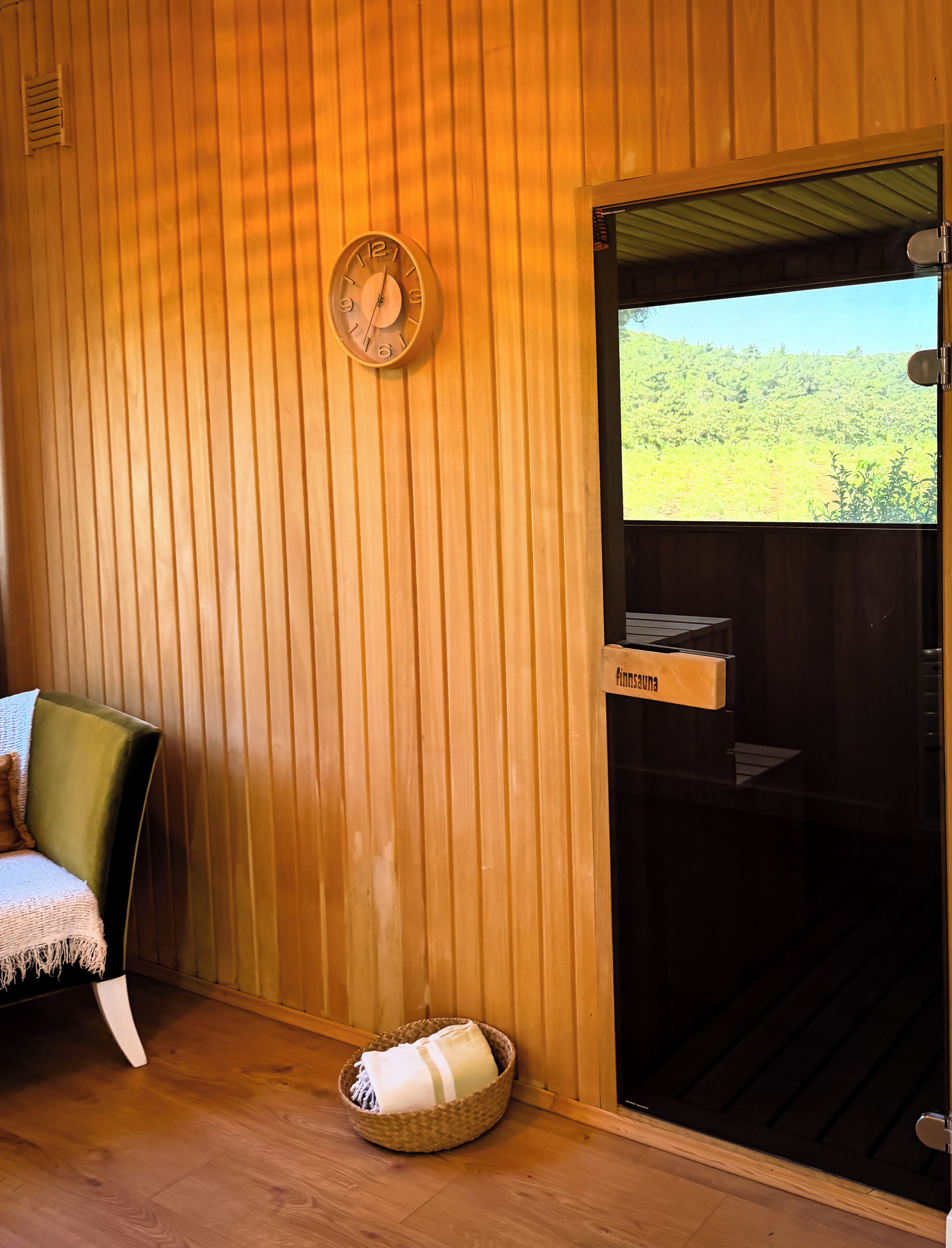 Un interior de madera con un reloj de pared, una puerta corredera que da a una vista exterior, una silla y una cesta en el suelo.