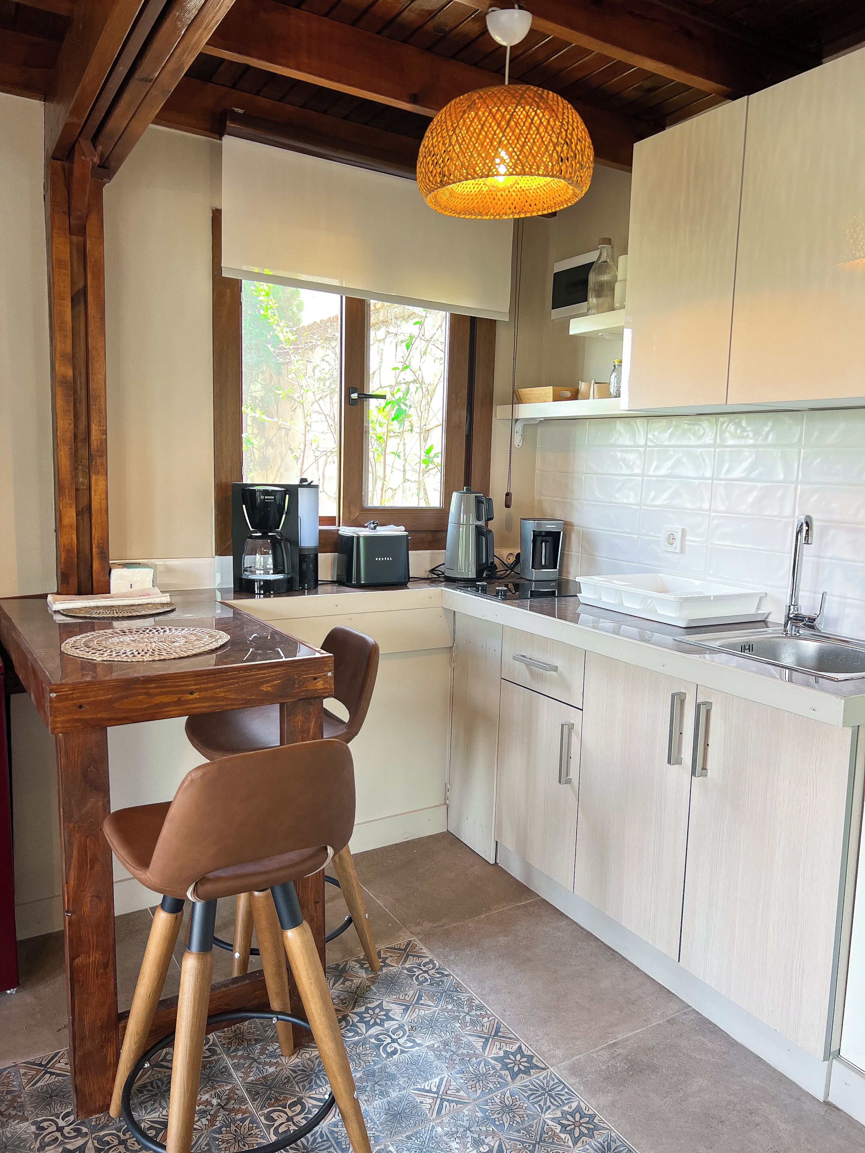 A kitchen with a wooden table, chairs, a hanging light fixture, and a window with a view of trees.