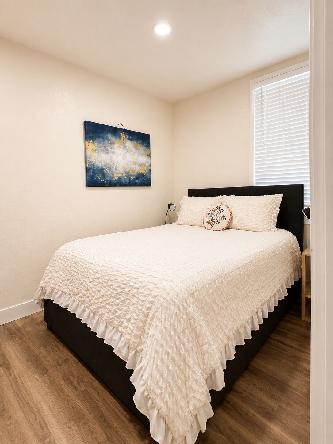 A bedroom with a bed, a wall-mounted painting, a bedside table, and a window with blinds.