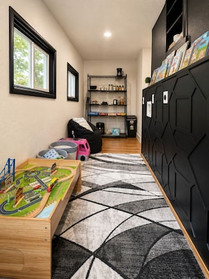 A room with a wooden play table, a black storage unit, a shelf with books, and a window.