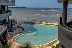 A pool with a curved edge, surrounded by a concrete deck and potted plants, overlooking a beach and ocean. - Oceanfront 4BR Duplex in Maiim Tankah w/Pool & Gym (Tulum)