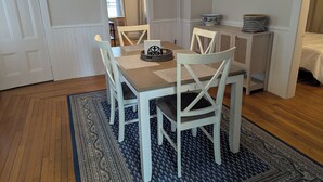 Spiseområde med bord, stole, et tæppe, et skab og en seng i baggrunden.