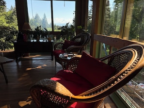 A room with wicker furniture, a red cushion, and a view of a forest and mountains through large windows.