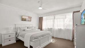 Un dormitorio con cama, mesitas de noche, ventilador de techo, ventana con cortinas y televisor.