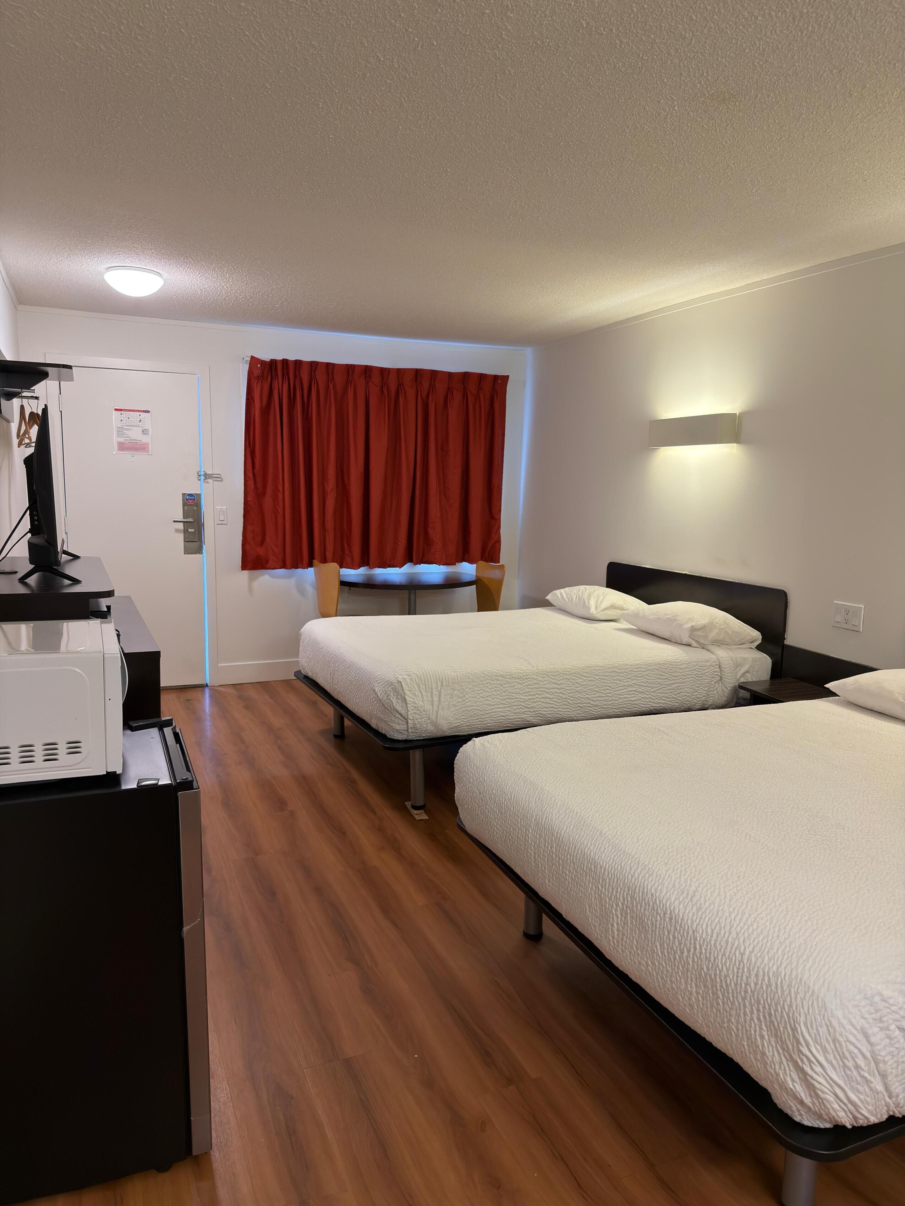 Habitación de hotel con dos camas, un escritorio con una computadora, una ventana con cortinas rojas y una lámpara fijada a la pared.