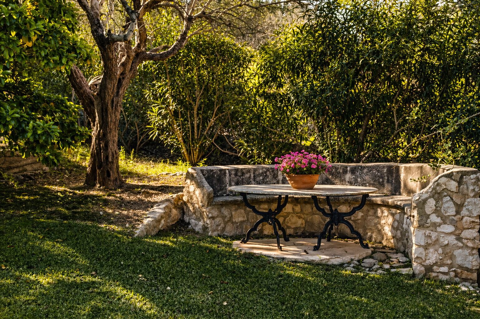 A stone bench with a table, surrounded by greenery and a tree.