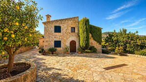 A stone building with a central arched doorway, a stone staircase leading to the entrance, and a well-maintained garden with various plants and a tall tree. - Can Gallardo d'en Fuster by Mallorca North Villas (Pollensa / Pollença)