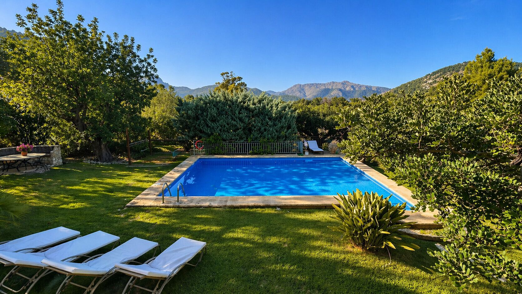 Une piscine rectangulaire bordée de pierres, entourée d’une pelouse verdoyante et d’arbres, avec une chaîne de montagnes en arrière-plan.