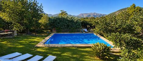 Une piscine rectangulaire bordée de pierres, entourée d’une pelouse verdoyante et d’arbres, avec une chaîne de montagnes en arrière-plan.