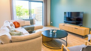 Ein modernes Wohnzimmer mit einer Couch, einem runden Couchtisch, einem Fernseher auf einem Holzschrank und einer Glastür zum Außenbereich.