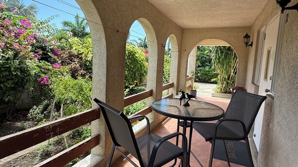 Une terrasse couverte avec une table ronde et des chaises, un banc, et une vue sur la verdure et les fleurs.