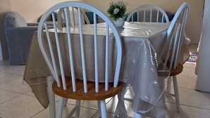Un espace repas avec une table recouverte d’une protection, entourée de chaises blanches à assise en bois.