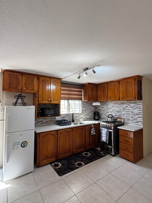 A kitchen with wooden cabinets, a white refrigerator, a microwave, a stove, and a window with blinds. - Affordable and spacious  2BR/2BA Close to Airport & South Coast (sleeps 4) (Oistins)