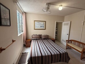 Una camera da letto con un letto, un comodino, una panca, una ventola a soffitto, un quadro incorniciato, una finestra con inferriate e una porta che dà su un armadio.