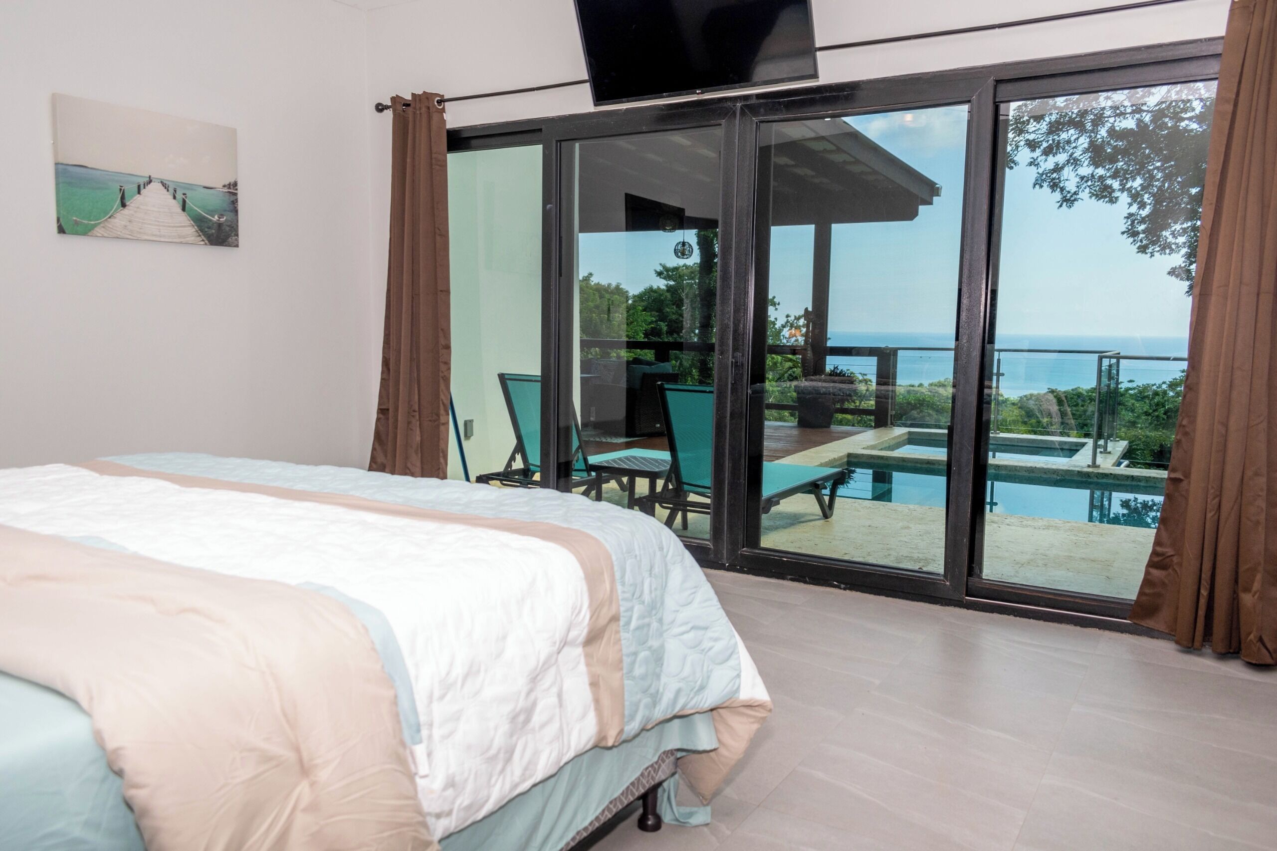 Ein Schlafzimmer mit Bett, Fernseher und einer Glasschiebetür, die auf einen Balkon mit Pool und Gartenmöbeln führt.