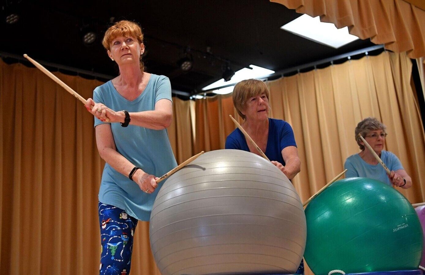 Trois personnes font de l’exercice avec des ballons et des bâtons dans une pièce où sont suspendues des rideaux.