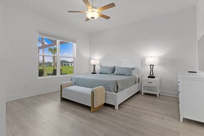A bedroom with a bed, bedside tables, a nightstand, a ceiling fan, and a window with a view of a green landscape. - Edenfield Golfview Retreat (Sumterville)