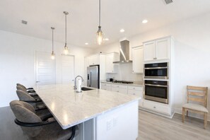 A modern kitchen with a central island, stainless steel appliances, pendant lighting, and a wooden chair. - Edenfield Golfview Retreat (Sumterville)