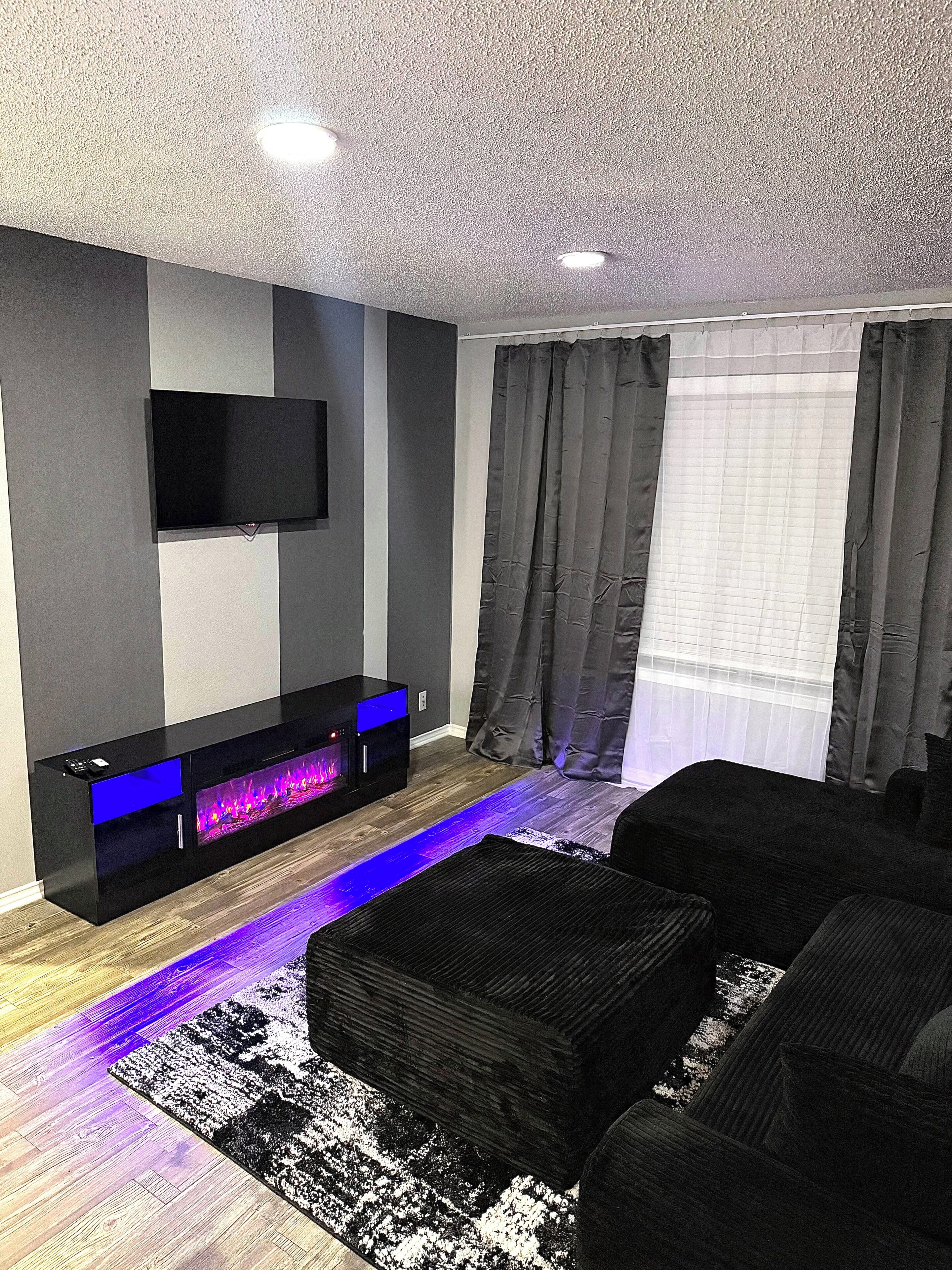 A modern living room with a flat-screen TV mounted on the wall, a built-in entertainment unit with a blue-lit display, a black sofa, and a patterned rug.