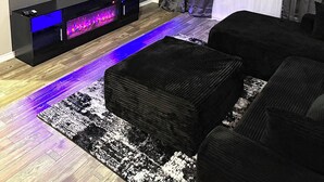 Una sala de estar moderna con un televisor de pantalla plana montado en la pared, un mueble empotrado con iluminación azul y un sofá negro, además de una alfombra estampada.