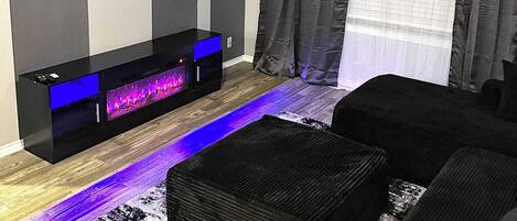 A modern living room with a flat-screen TV mounted on the wall, a built-in entertainment unit with a blue-lit display, a black sofa, and a patterned rug.