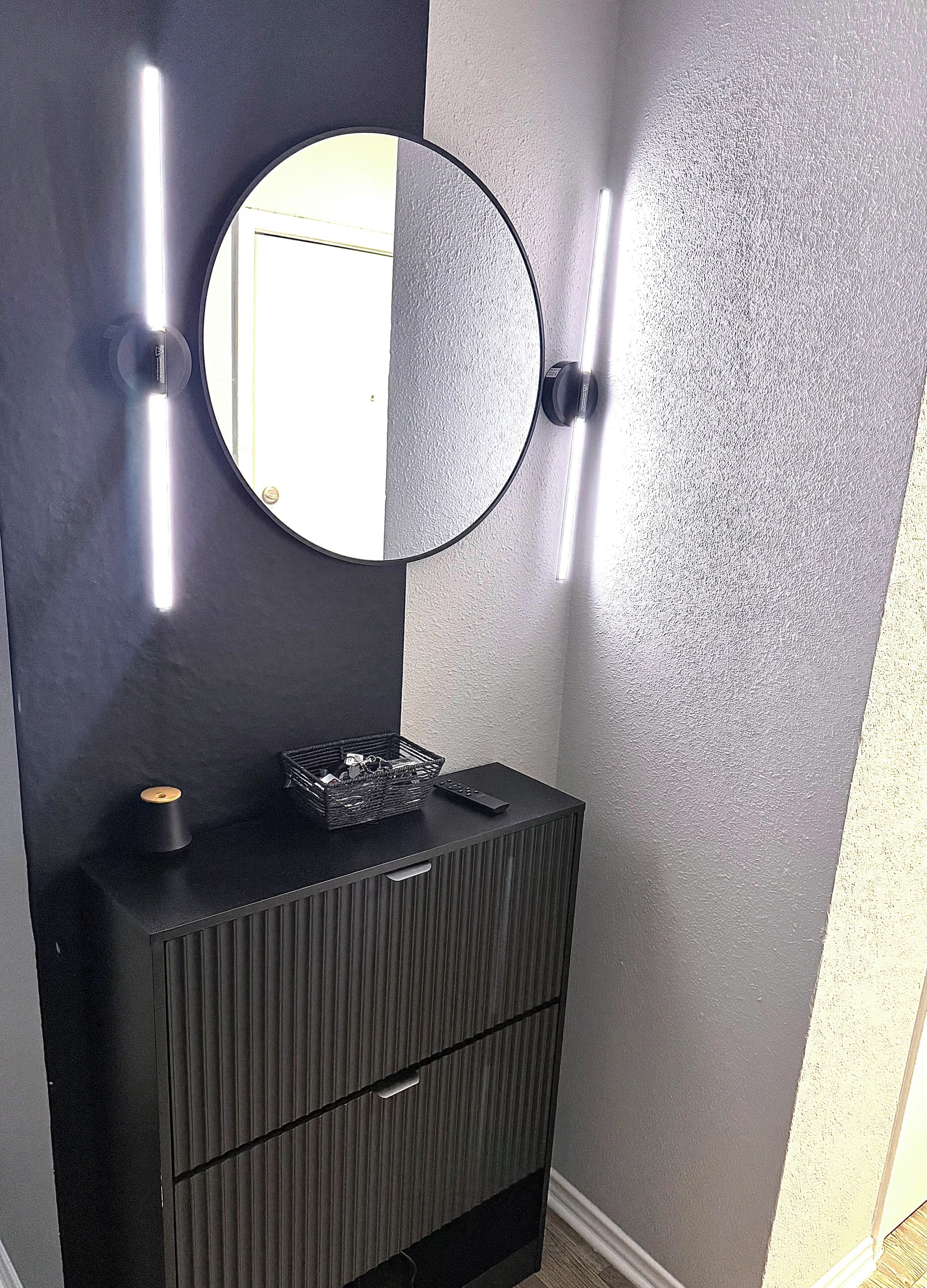 Una habitació amb un armari fosc, un mirall rodó i dues làmpades fixades a la paret.