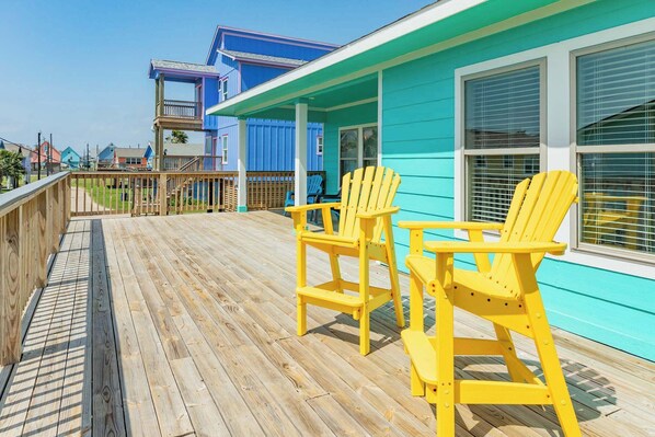 Um deck de madeira com cadeiras Adirondack amarelas, um edifício azul com detalhes brancos e uma cerca de madeira.