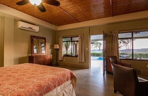 A bedroom with a bed, a ceiling fan, a window with curtains, a chair, and a view of the outdoors.