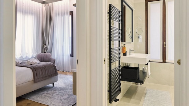 Un dormitorio con cama, mesita de noche, espejo y ventana con cortinas.
