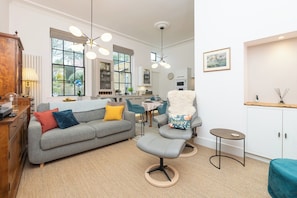 Un salon moderne avec un canapé gris, un pouf et un fauteuil agrémenté d’un coussin moelleux. On y trouve une petite table ronde et un meuble avec un plan de travail en bois.