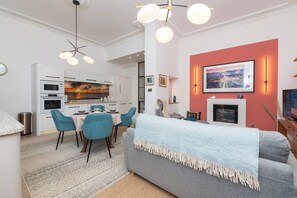 Une cuisine moderne avec des meubles blancs, un coin repas avec des chaises bleues et un salon avec un canapé gris.