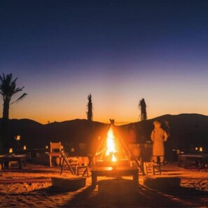 A campfire at dusk with a person standing nearby, surrounded by sand and sparse vegetation. - Sunbright luxury camp (Merzouga)