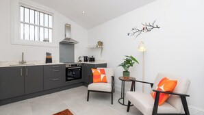 A modern kitchen with grey cabinets, a white countertop, and a window. There are two chairs with orange cushions, a small round table, and a floor lamp. - One Bedroom Retreat in Prime Location - Pass the Keys (Bath)