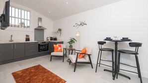 A modern kitchen with grey cabinets, a built-in oven, and a large window. There are two chairs and a table with stools, a rug, and a wall-mounted light fixture. - One Bedroom Retreat in Prime Location - Pass the Keys (Bath)