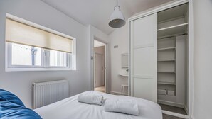 A bedroom with a bed, a window with blinds, a radiator, a white ceiling light, and a white wardrobe.