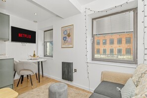 A modern living room with a flat-screen TV, a small table with a bottle, a grey chair, a grey ottoman, and a window with blinds. - Dreamy Leeds Centre Retreat – Ideal for Couples - Pass the Keys (Leeds)