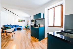 A modern kitchen with a central island, a dining area with a blue sofa, and a large window providing natural light. - Heritage De Amicis old Town (Rovinj)