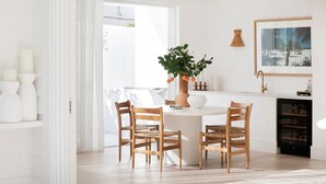 Een lichte kamer met een ronde tafel, vier houten stoelen, een potplant, een vaas, een lamp en een lijst met een afbeelding aan de muur.