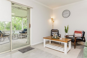 Quarto com porta de vidro que dá para área externa, mesa de centro de madeira, relógio na parede e uma cadeira com cobertor.