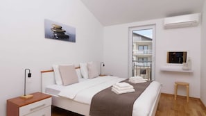 Ein Schlafzimmer mit Bett, Nachttischen, Fenster, Balkon und einer Wand-Klimaanlage.