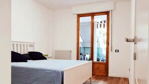 Quarto com cama, piso de madeira, radiador branco e porta de correr para uma varanda.